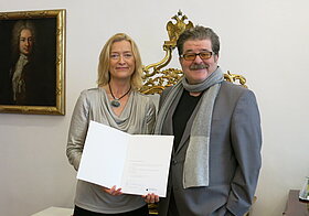 Zwei Personen posieren mit Urkunde vor Gemälde.