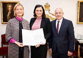 Zwei Personen in eleganter Kleidung halten ein Dokument. Rechts eine Person im Anzug.