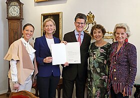 Fünf Personen halten ein Dokument. Im Hintergrund ein Gemälde und eine Standuhr. 