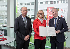 Drei Personen, zwei davon halten Urkunde.