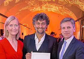 Drei Personen in eleganter Kleidung posieren lächelnd mit Urkunde.